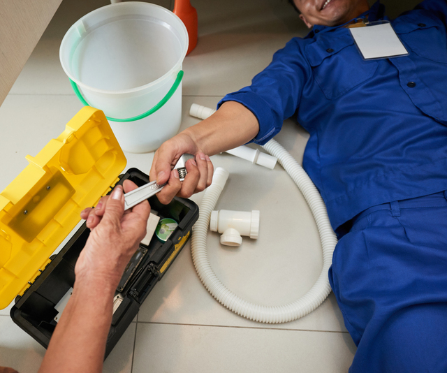 Senior Woman Passing Wrench To Plumber Who Is Lying On The Floor