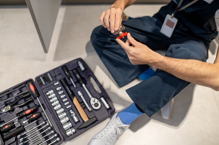 Ball Valve. Top View Of Man In Blue Overalls With Badge And White Sneakers With Ball Valve In Hands Sitting On Floor Near Set Of Tools, Without Face