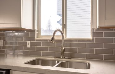 Polished kitchen countertop with double basin stainless steel sink and faucet. Wood cabinets and window are installed above the gray tiled wall.