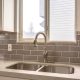 Polished kitchen countertop with double basin stainless steel sink and faucet. Wood cabinets and window are installed above the gray tiled wall.