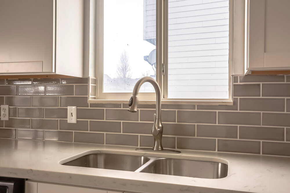 Polished kitchen countertop with double basin stainless steel sink and faucet. Wood cabinets and window are installed above the gray tiled wall.
