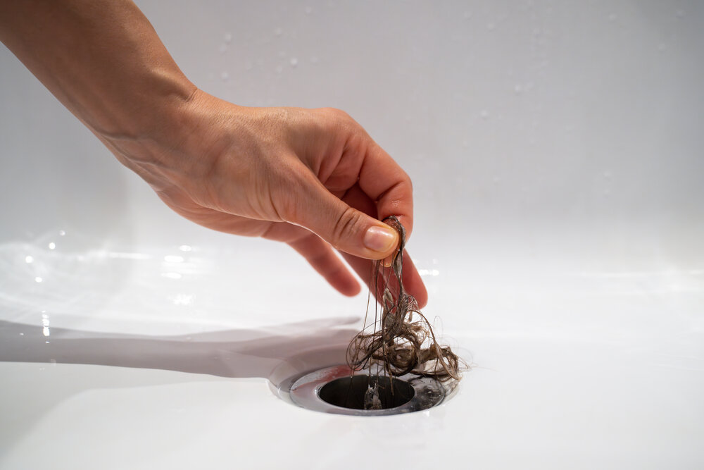 Water drain hole in bath is clogged with hair clump in bathroom at home. Female cleaning sewer trap taking hairs, hand closeup. Bath plug hole blocked with dirt. Problem with shower sewerage.