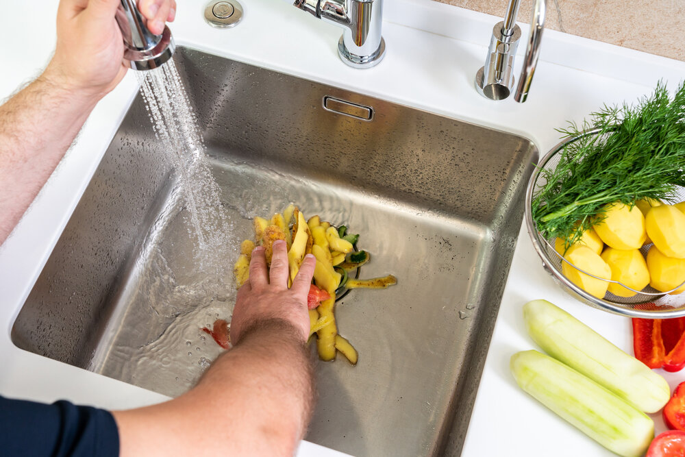 A hand shoves food waste into a disposer hole in the kitchen sink.
