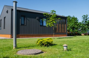 Residential utility infrastructure near cottage, featuring sewer manhole cover, sewer cleanout pipe and exterior utility pole installed in landscaped lawn beside modern house