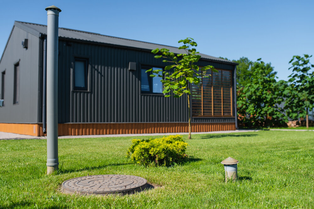 Residential utility infrastructure near cottage, featuring sewer manhole cover, sewer cleanout pipe and exterior utility pole installed in landscaped lawn beside modern house